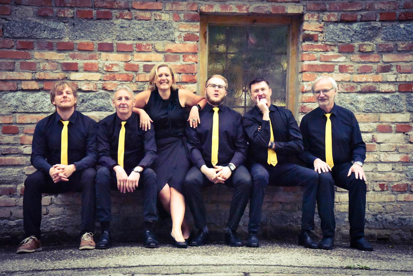 Die Band in Schwarz mit gelben Krawatten vor einer Ziegelmauer